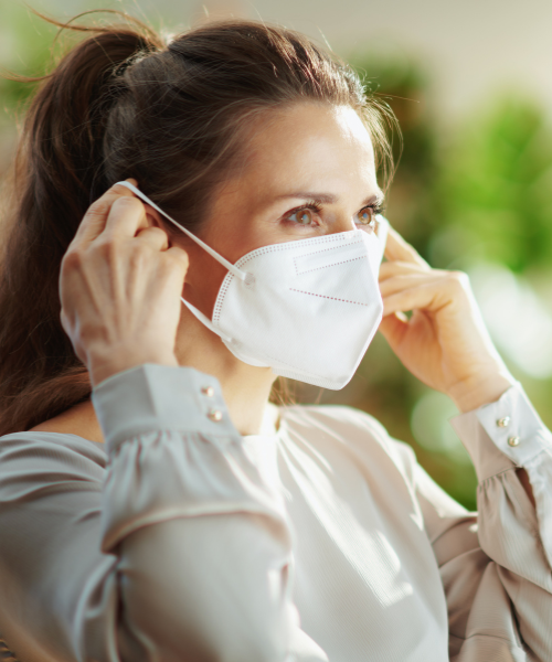 Woman putting on a white N95 face mask indoors, with greenery in the background. For a visual tip on respiratory health in high pollution.