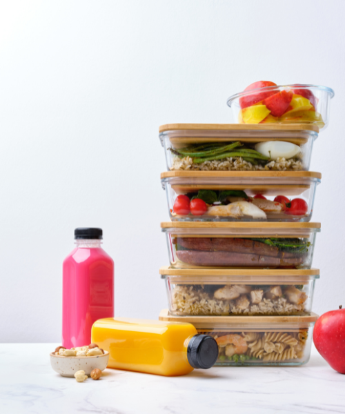 A neatly stacked set of meal prep containers filled with healthy meals, alongside fruit juice and nuts on a white background.